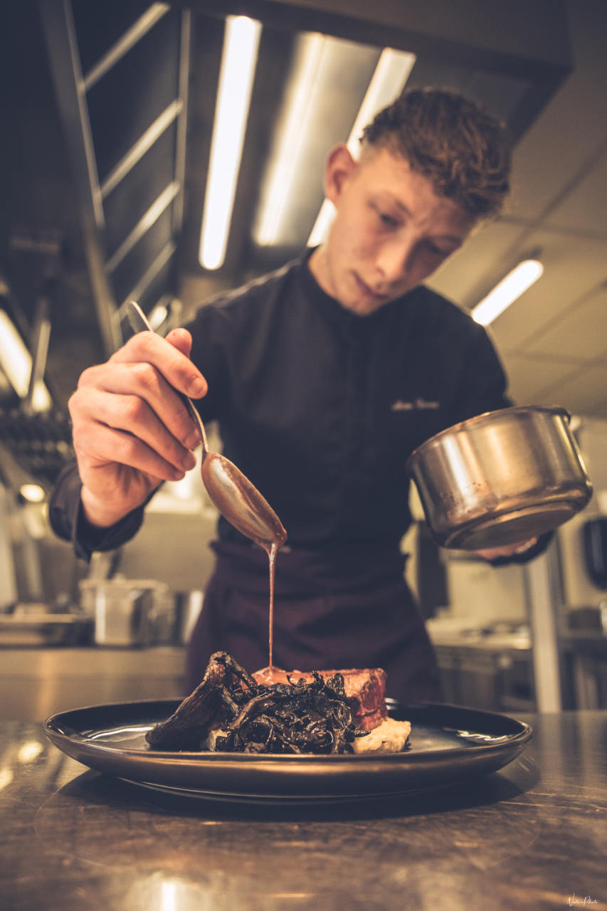 Un chef nappe de sauce une assiette en cuisine, casseroles au premier plan