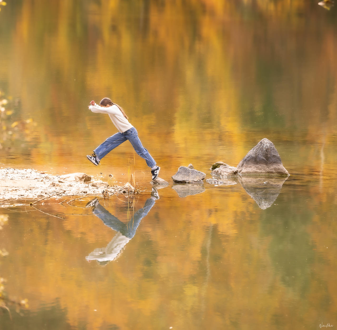 Un garçon saute de pierre en pierre, son reflet dans l’eau dorée d’un lac
