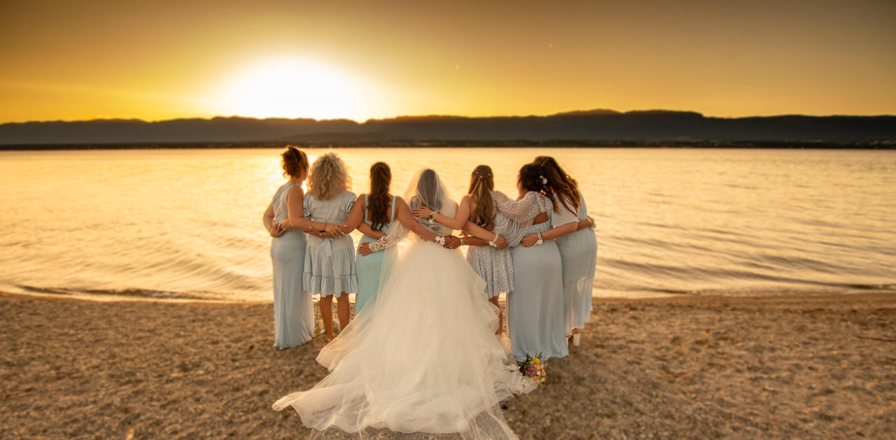 La mariée et ses amies, de dos, enlacées au bord de l’eau au coucher du soleil