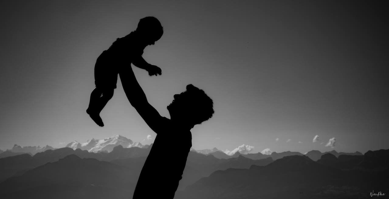 Silhouette d’un père qui lance son bébé en l’air, montagnes au loin, en noir et blanc