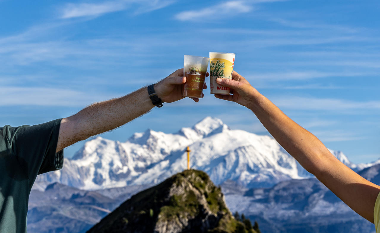 Deux mains trinquent devant un massif enneigé, sous un ciel bleu